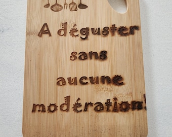 May include: A rectangular bamboo cutting board with a rounded top corner and a hanging hole. The board features a laser-engraved illustration of five kitchen utensils and the French text: A déguster sans aucune modération!
