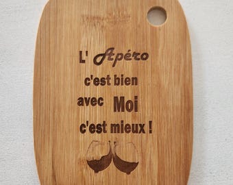 May include: A rectangular bamboo cutting board with rounded corners and a circular hole at the top right. The board features laser-engraved text that reads L' Apéro c'est bien avec Moi c'est mieux! and an illustration of two wine glasses.