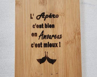 May include: A rectangular bamboo cutting board with rounded corners featuring a laser-engraved design. The text reads L' Apéro c'est bien en Amoureux c'est mieux! above a graphic of two wine glasses clinking together in a toast.
