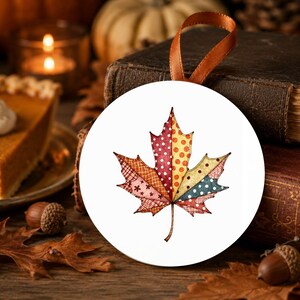 May include: A round white ceramic ornament featuring a colourful maple leaf illustration divided into segments with various patterns including polka dots, cross-hatching, and floral motifs in shades of red, orange, yellow, and blue with a brown ribbon.