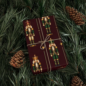 May include: A rectangular gift box wrapped in dark maroon paper featuring a repeating pattern of nutcracker figures in green and gold uniforms, tied with thin tan twine and accented with small gold star icons across the surface.
