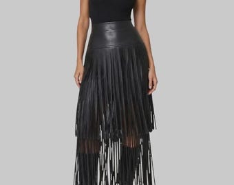 Op de afbeelding: Een zwarte mouwloze top met opstaande kraag gecombineerd met een zwarte midi-rok met hoge taille, voorzien van meerdere lagen lange, dunne franjes. De outfit wordt gecompleteerd door zwarte enkellaarsjes met een spitse neus en naaldhak.