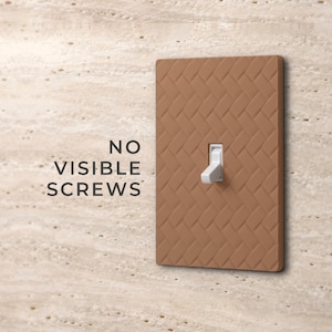 May include: A rectangular light switch cover plate with a brown, textured herringbone pattern and a white toggle switch in the center. The text NO VISIBLE SCREWS is printed to the left of the switch plate on the wall surface.