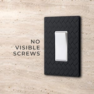 May include: A black light switch wall plate with a textured herringbone pattern and a white rocker switch in the center. The text NO VISIBLE SCREWS is printed to the left of the plate, which is mounted on a light beige stone wall.