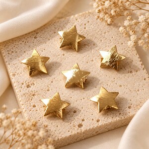 May include: Six gold-toned star-shaped hair clips with a brushed metal texture. Each star features five points and a polished finish that catches the light, arranged in a cluster on a flat surface to display their uniform design and metallic sheen.