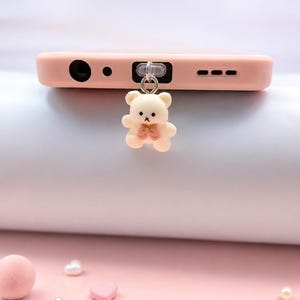 May include: A light pink smartphone case featuring a small, cream-colored bear charm attached to the bottom edge. The bear has a tiny pink bow at its neck and is suspended by a silver-toned metal ring connected to a clear plastic loop on the case.