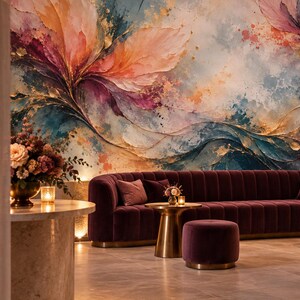 May include: A long, channel-tufted velvet sofa in deep plum, paired with two matching cylindrical ottomans and two gold-toned side tables. The wall behind features a large-scale mural with abstract, flowing brushstrokes in pink, blue, and gold.