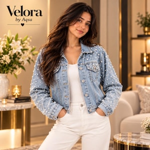 Könnte beinhalten: Eine hellblaue Jeansjacke, die mit verstreuten weißen Perlen auf der Vorderseite und den Ärmeln verziert ist, mit klassischem Kragen, Metallknöpfen und zwei Brusttaschen. Der Text Velora by Aqsa ist in der oberen linken Ecke zu sehen.