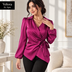 Könnte beinhalten: Eine magentafarbene Wickelbluse aus Satin mit langen Ärmeln, gerafften Bündchen, einem V-Ausschnitt und einem seitlichen Bindegürtel. Das Kleidungsstück trägt das Etikett Velora by Aqsa. Der Stoff hat eine glatte, reflektierende Oberfläche.