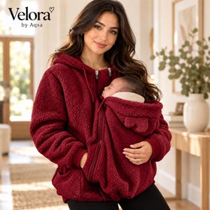 Könnte beinhalten: Ein dunkelroter, langärmeliger Kapuzenpullover mit Reißverschluss aus strukturiertem Fleece-Material. Das Kleidungsstück verfügt über eine Kapuze mit Kordelzug und eine spezielle Fronttasche für einen Säugling. Der Markenname Velora by Aqsa ist schwarz aufgedruckt.