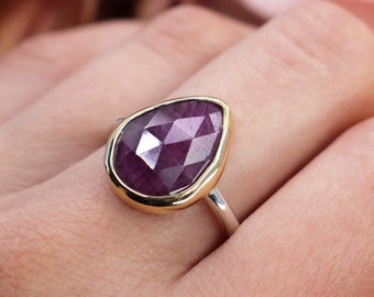 May include: A teardrop-shaped ring with a faceted purple gemstone set in a gold bezel. The ring is on a silver band and is worn on a person's finger.