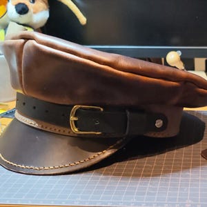 May include: A brown leather military-style cap featuring a black leather strap across the front, secured with a gold-toned metal buckle. The cap has a structured visor with visible tan stitching along the edge and a smooth, dark brown crown.