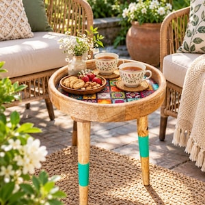 May include: A round wooden side table featuring a colorful mosaic tile tabletop with geometric patterns. The table has three light-colored wooden legs, each accented with a bright turquoise painted band near the bottom. The surface holds two floral tea cups, a small vase with white flowers, and a wooden bowl filled with strawberries and cookies.