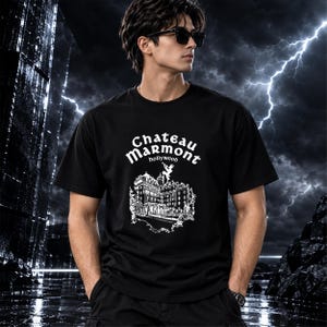 May include: A black short-sleeved t-shirt featuring a white graphic print of the Chateau Marmont building in Hollywood. The text reads Chateau Marmont hollywood, with a small figure of a person playing a trumpet positioned above the building.