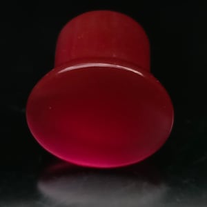 May include: A solid, deep red ear plug with a smooth, glossy finish. The jewelry features a wide, circular front face that tapers into a narrower cylindrical body, designed for insertion into a stretched ear piercing, presented against a dark background.