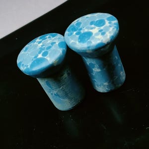 May include: Two cylindrical ear plugs featuring a flat, circular top surface with a mottled pattern of light blue, medium blue, and white circular spots. The sides of the plugs transition from a smooth blue-spotted top to a textured, darker base.