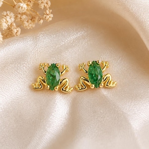 Pode incluir: Um par de brincos de pino dourados em forma de sapo, cada um com uma pedra preciosa verde de corte marquise como corpo. O design de metal inclui pernas e pés detalhados que se estendem dos lados da pedra verde vibrante.