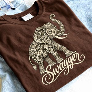 May include: A brown t-shirt featuring a large, cream-colored graphic of an elephant decorated with intricate floral and geometric patterns. The word "Swagger" is printed in a cursive, cream-colored script font below the elephant illustration.