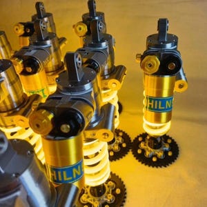 May include: A collection of multiple Öhlins suspension shock absorbers featuring gold-coloured cylindrical bodies with blue labels, black metal mounting heads, and white coiled springs. Each unit includes black circular gear-like base plates.