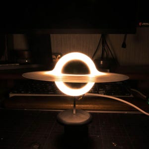 Könnte beinhalten: Eine Schwarzes-Loch-Lampe mit einem leuchtenden, kreisförmigen Lichtring, der eine flache, durchscheinende Scheibe beleuchtet. Die Struktur wird von einem dünnen, vertikalen Metallstab auf einem soliden, dunklen, zylindrischen Sockel getragen.