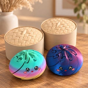 Könnte beinhalten: Zwei runde, bewegliche Fidget-Spielzeuge in Form von gedämpften Brötchen, eines mit Regenbogenverlauf und das andere mit metallisch blauer und violetter Oberfläche. Beide haben ein aufgedrucktes Cartoon-Gesicht mit schwarzen Augen und roten Wangen und stehen vor zwei beigen Bambusdampfkörben.