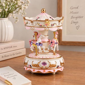 May include: A decorative carousel music box featuring white horses with pink manes and tails, accented with gold poles. The structure is white and pink with floral details, gold trim, and a central purple gem on the base, topped with a gold finial.