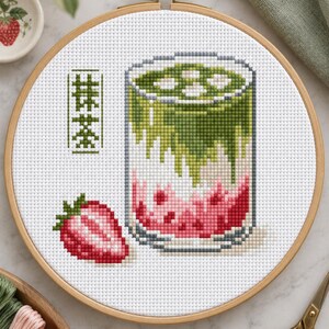 Könnte beinhalten: Ein Kreuzstich-Stickprojekt in einem runden Holzrahmen, das ein Glas mit einem geschichteten Matcha-Erdbeer-Getränk zeigt. Das Design enthält eine leuchtend rote Erdbeere und japanische Kanji-Schriftzeichen für Matcha auf weißem Aida-Stoff.