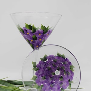 May include: Two clear martini glasses with hand-painted purple hydrangea flowers and green leaves. The flowers have yellow centers. One glass is upright, the other is tilted, showing the floral design inside. Green decorative grass is in the foreground.