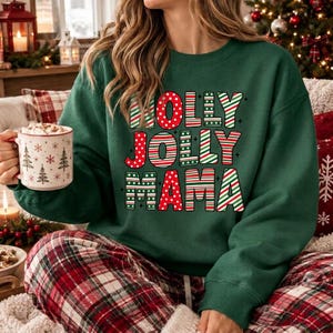 May include: A dark green crewneck sweatshirt featuring the text HOLLY JOLLY MAMA in a bold, multi-colored font with red polka dots and green and white diagonal stripes. The sweatshirt is paired with red and white plaid pajama pants.