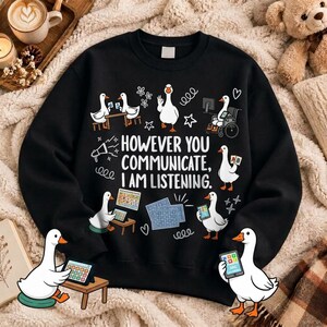 Könnte beinhalten: Ein schwarzes Sweatshirt mit Rundhalsausschnitt und einem Grafikdruck von weißen Gänsen, die verschiedene Kommunikationsgeräte benutzen. Der Text lautet: "HOWEVER YOU COMMUNICATE, I AM LISTENING." Das Design zeigt Gänse mit Tablets, einem Rollstuhl und Diagrammen.