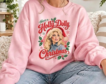 Op de afbeelding: Een lichtroze sweater met ronde hals en een grafische print van een blonde vrouw met een rode kerstmuts. De tekst luidt Have a Holly Dolly Christmas, omringd door rode hulstbessen, groene bladeren en kleine rode sneeuwvlokken op de voorzijde.