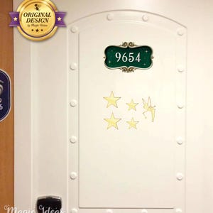 Può includere: Una porta bianca con una targa rettangolare verde scuro che riporta il numero 9654 in bianco. Sotto la targa ci sono quattro stelle dorate e la sagoma dorata di una fata con le ali. La porta presenta un design a pannelli in rilievo.