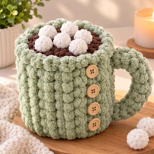 Puede incluir: Una funda de ganchillo para taza en color verde salvia con un patrón texturizado de puntos de burbuja y cuatro botones de madera clara en el lateral. La parte superior de la taza está rellena de líquido marrón oscuro y malvaviscos blancos.