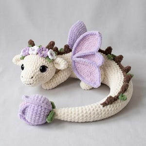 May include: A cream-colored crochet dragon plush toy featuring lavender wings and a matching lavender flower bud at the end of its tail. The dragon has dark brown horns and back spikes, green leaf accents, and a floral crown of white and lavender flowers.