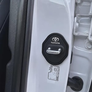 May include: A black octagonal Toyota door latch cover installed on a white vehicle door frame. The cover features the white Toyota logo and text. Below the latch, a small white rectangular sticker with black text and diagrams is visible.