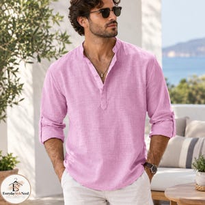 May include: A light pink long-sleeve shirt with a mandarin collar and a partial button-down placket. The shirt features a textured fabric, rolled-up sleeves, and a curved hemline. A silver chain necklace is worn around the neck with the shirt.