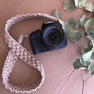 May include: A black Canon digital camera with an 18-55mm lens attached to a beige macrame camera strap featuring a knotted cord pattern. The strap is looped in a figure-eight shape, and a green eucalyptus branch is positioned to the right side.