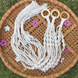 Puede incluir: Tres colgadores de plantas de macramé en color blanco roto con patrones de cordón de algodón anudado y anillos de madera clara en la parte superior. Los colgadores tienen flecos largos y sueltos y están sobre una bandeja redonda de bambú.