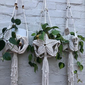 Può includere: Tre portapiante in macramè color bianco sporco con un design a forma di stella, anelli in legno nella parte superiore e lunghe nappe annodate sul fondo. Ogni supporto è decorato con piccole perline di legno e contiene una pianta verde.