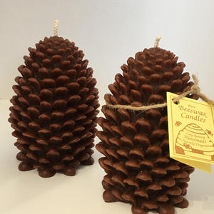 Może przedstawiać: Dwie brązowe świece z wosku pszczelego w kształcie szyszek. Każda świeca ma knot i etykietę z napisem "Pure Beeswax Candles, Long Burning, Handmade, Sweetly Fragrant."