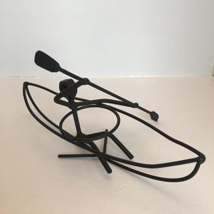 Puede incluir: Escultura de metal negro de una persona en una canoa con un remo. La escultura está sobre un fondo blanco.