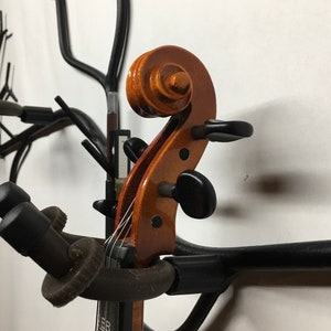 Könnte beinhalten: Eine Holzvioline mit brauner Oberfläche und schwarzen Stimmwirbeln ist auf einem schwarzen Metallständer montiert. Die Violine befindet sich in vertikaler Position mit der Schnecke zum Betrachter.