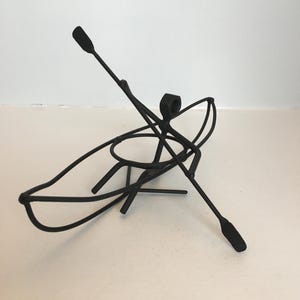 Puede incluir: Escultura de metal negro de una persona en una canoa con dos remos. La escultura es una representación caprichosa y abstracta de una persona remando en una canoa.