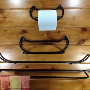 May include: Three black metal canoe-shaped shelves mounted on a wood wall. The top shelf is a toilet paper holder, the middle shelf is empty, and the bottom shelf has a red and green patterned towel hanging on it.