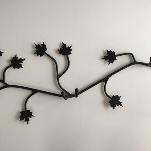 Könnte beinhalten: Schwarzer Wandhaken aus Metall in Form eines Astes mit Blättern. Der Haken hat mehrere Äste und Blätter und ist dafür ausgelegt, Gegenstände wie Mäntel, Hüte oder Taschen zu halten.