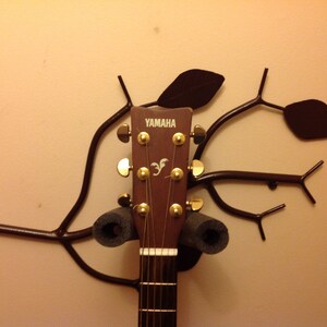 Könnte beinhalten: Eine braune Akustikgitarre mit goldenen Stimmwirbeln ist an einem schwarzen Metallwandhalter in Form eines Astes mit Blättern befestigt. Die Gitarre trägt den Schriftzug "YAMAHA" auf dem Kopf.