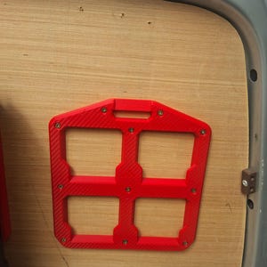 May include: A bright red, rectangular storage rack with a four-grid open design and a textured, carbon-fibre-like finish. The frame is secured to a light-coloured wooden panel with eight visible metal screws positioned at the corners and joints.