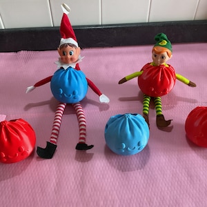 May include: Two elf dolls with pointed hats and striped legs, one wearing a red hat and the other a green hat. They are accompanied by four spherical, pleated plastic containers in red and blue, each featuring a small embossed smiling face.