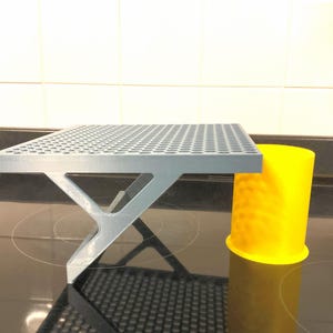 May include: A silver-coloured shelf with a perforated top surface and a triangular support bracket, positioned next to a bright yellow cylindrical container with a textured exterior surface featuring a repeating pattern of small circular indentations.