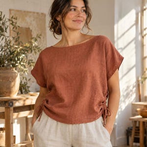 Puede incluir: Una blusa de manga corta color óxido con un amplio escote barco y cordones ajustables en ambos lados del dobladillo. La prenda presenta un acabado de tela texturizada y un corte holgado, combinada con pantalones claros en un tono neutro.